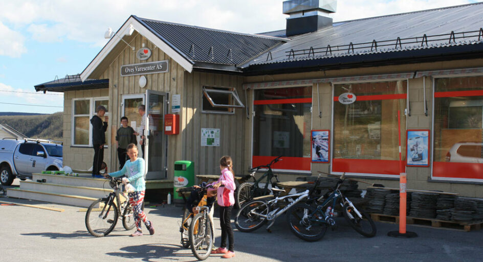 To barn med sykkel og tre voksne utenfor butikk. Foto.