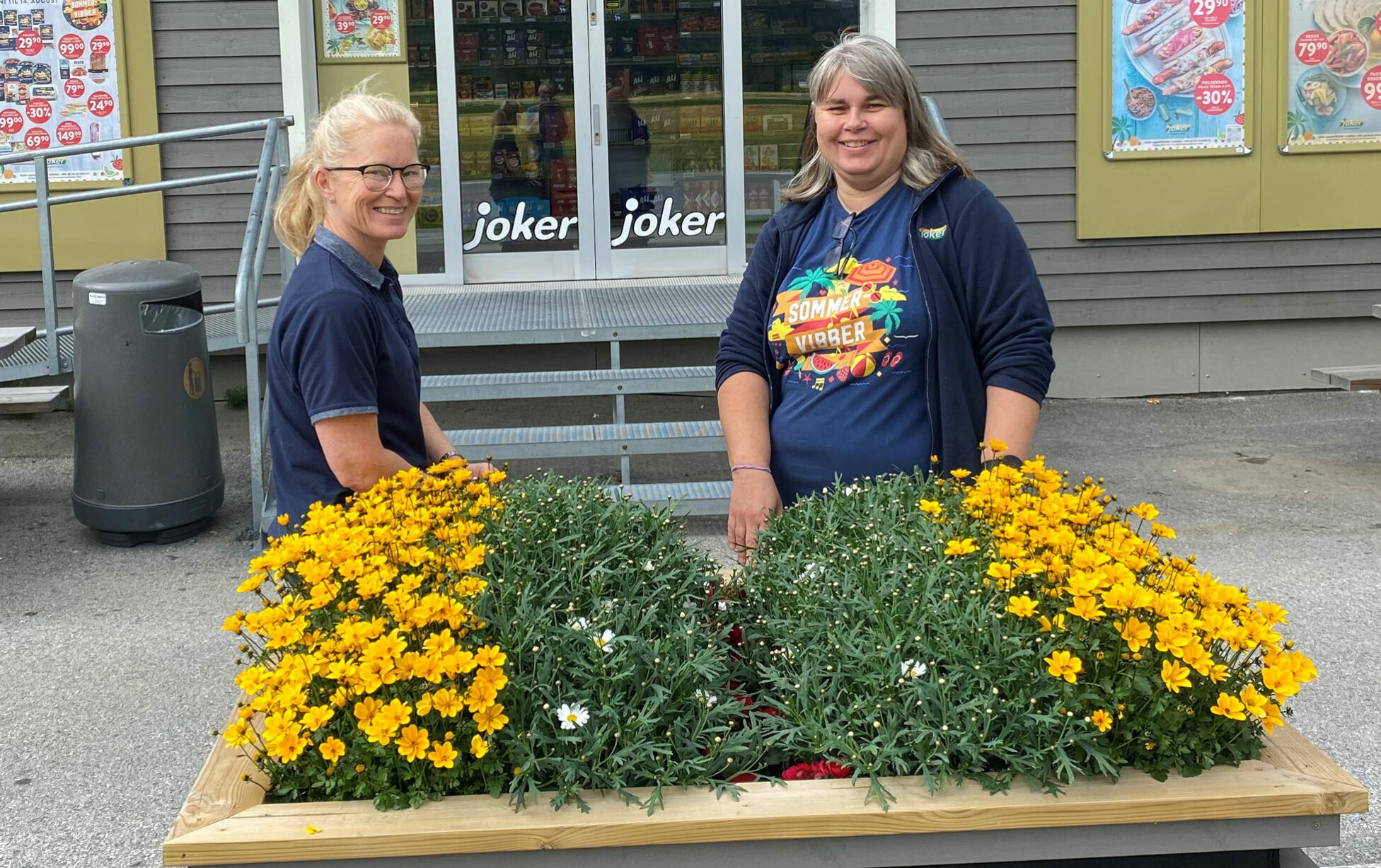 To personar framfor blomsterkasse utanfor inngangen til butikk. Foto.