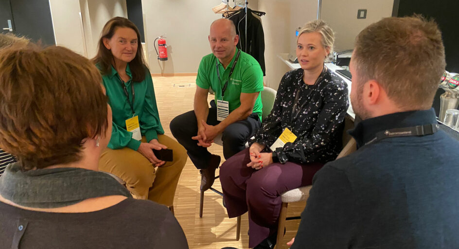 4 personer sitter på høye krakker og snakker sammen. Foto.