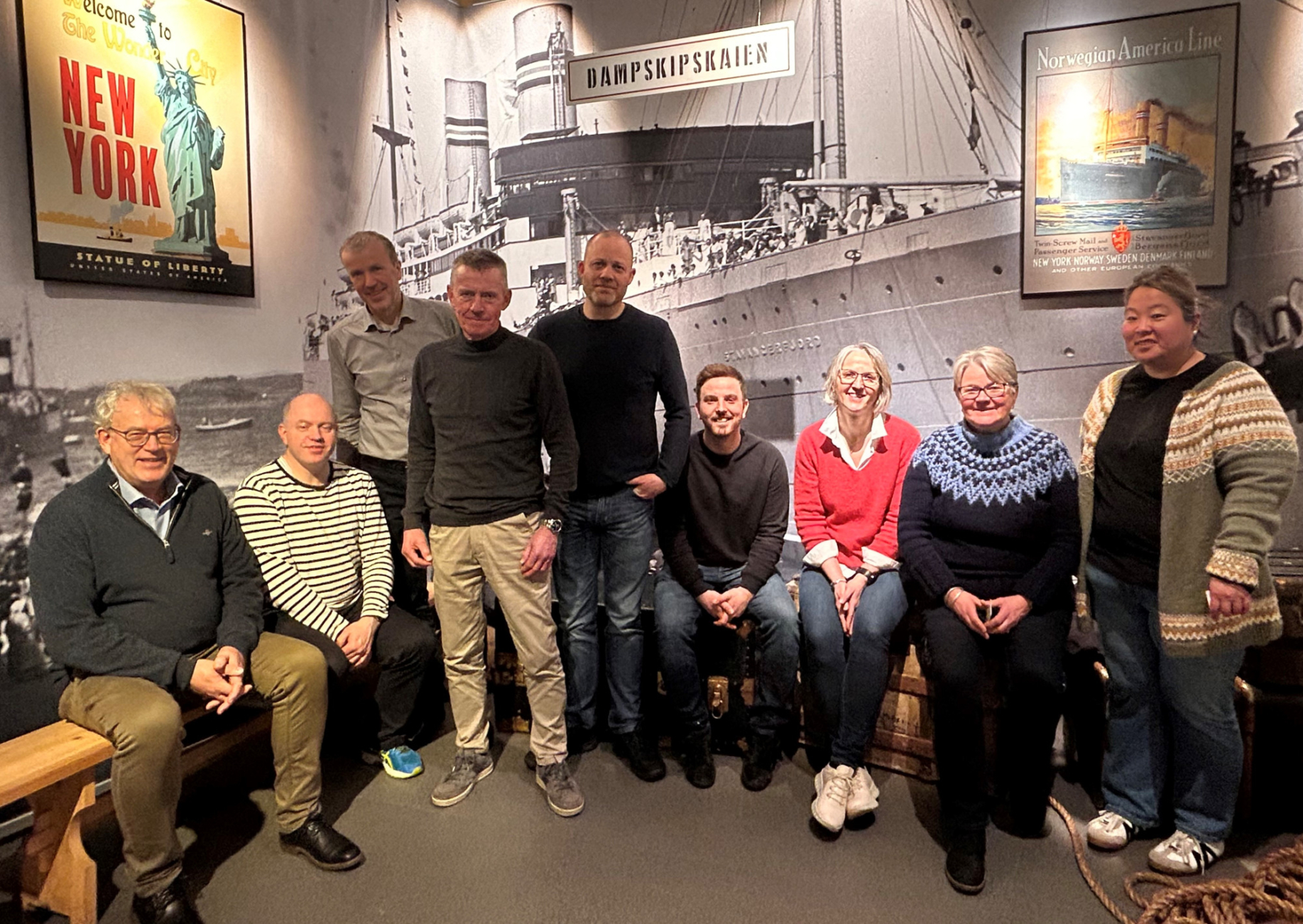 Foto av en gruppe på ni personer foran eit stort bilete av eit dampskip